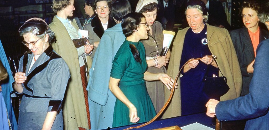 Members at a display table, examining leatherwork; one holds up a tooled belt. From the Society's photograph archive, undated.