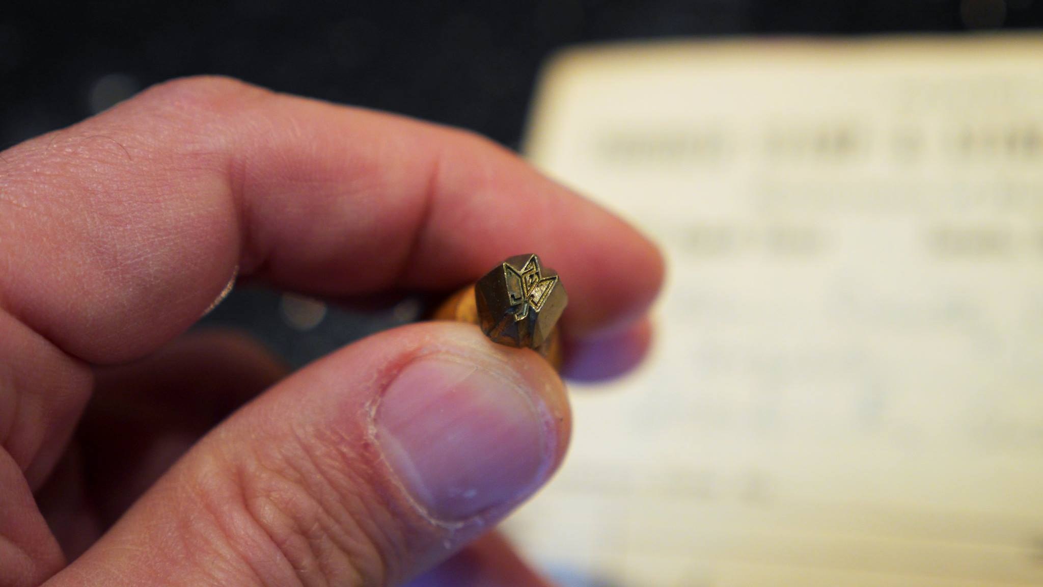 A stamp's brass die held between finger and thumb, showing the monogram.