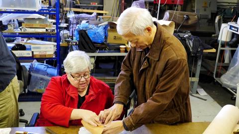A man in a brown jacket helps a woman fold leather around a box form at the workshop table
