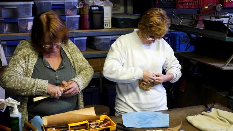 Two women work leather around box forms at the table, tools and supplies spread in front
