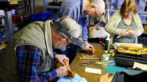 A man in a headband magnifier works leather around a form while others work behind him
