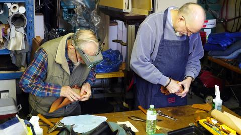 Two men in the workshop stretch leather over box forms, one wearing a headband magnifier