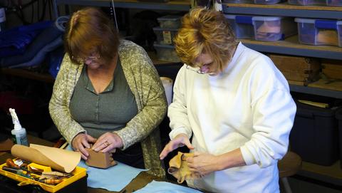 Two women work leather around small box forms at a table with a yellow toolbox