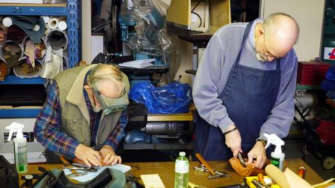 A man in a headband magnifier and a man in an apron each work leather over box forms