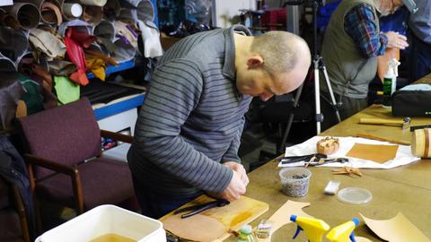 A man bends over the table pulling leather over a box form, cut leather and tools around him