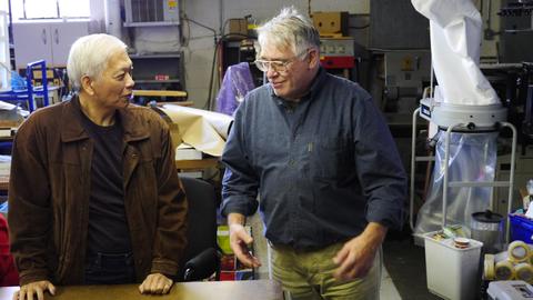 Two men talk beside a workshop table, a dust collector and supplies behind them