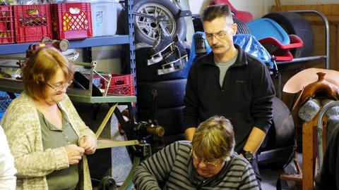 A woman holds a leather strip as a man watches and another member works at the bench