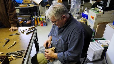 A man works leather over a box form in his lap at the end of a workbench