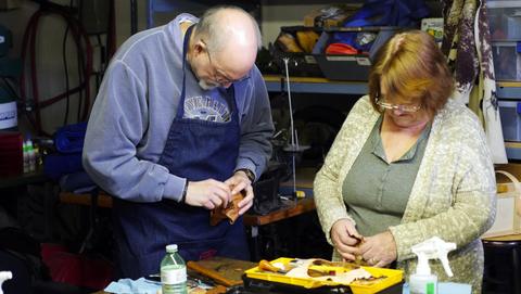 A man in an apron and a woman each shape leather over box forms at the workbench