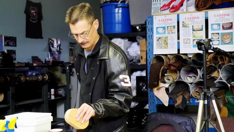 A man in a leather jacket holds a covered box form near a tripod and lasting box posters