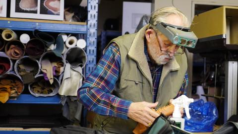 A man in a headband magnifier works leather over a form at a bench, rolled leather on shelves behind