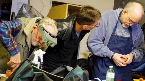 Three men lean in over the table, one in a headband magnifier, working on a lasted box