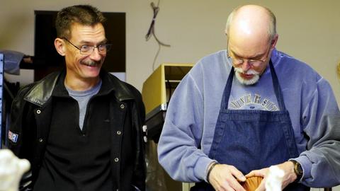 A smiling man in a leather jacket stands beside a man in an apron working on a lasted box