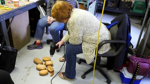 A seated woman uses a heat gun on lasted box forms laid out on the workshop floor