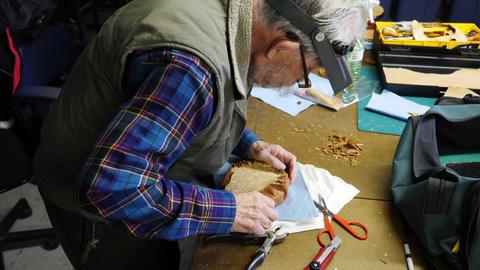 A man in a headband magnifier trims leather around a form, red-handled scissors beside him