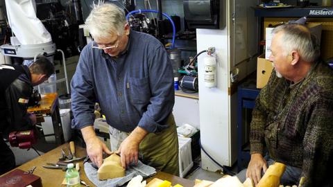A man stretches leather over a shaped form on a towel while another man watches from the side