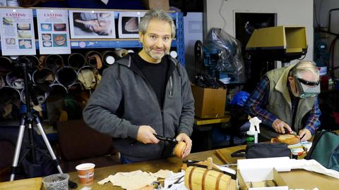 A smiling man holds lasting pliers at the table while a man in a headband magnifier works behind
