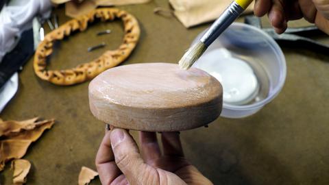 A brush spreads white glue on the top of a leather-covered oval form held in a hand