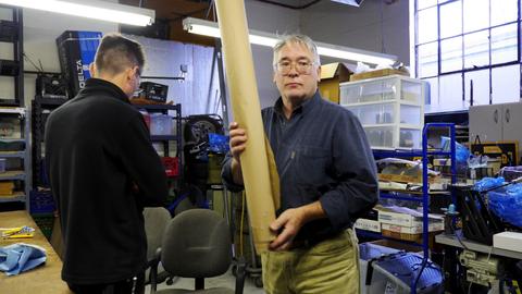 A man carries a tall roll of leather past another man in the workshop