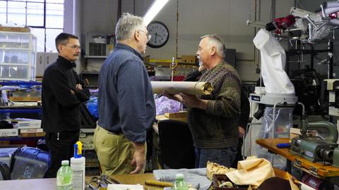 Three men talk in the workshop, one holding a roll of leather
